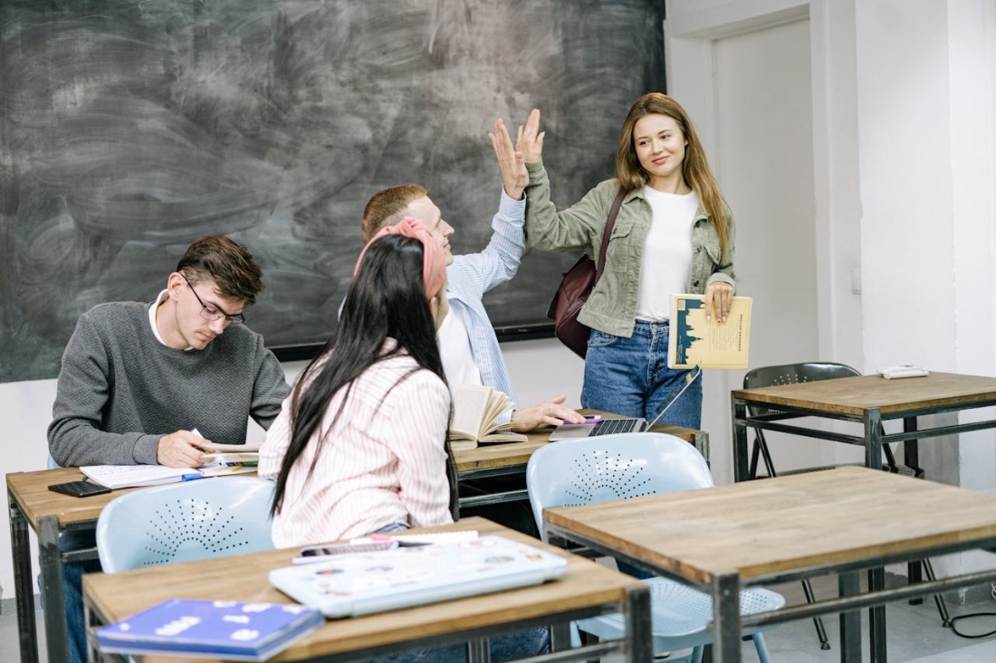 Studierende in einem Klassenzimmer, eine Person hebt die Hand, vorne steht eine junge Frau mit Buch.