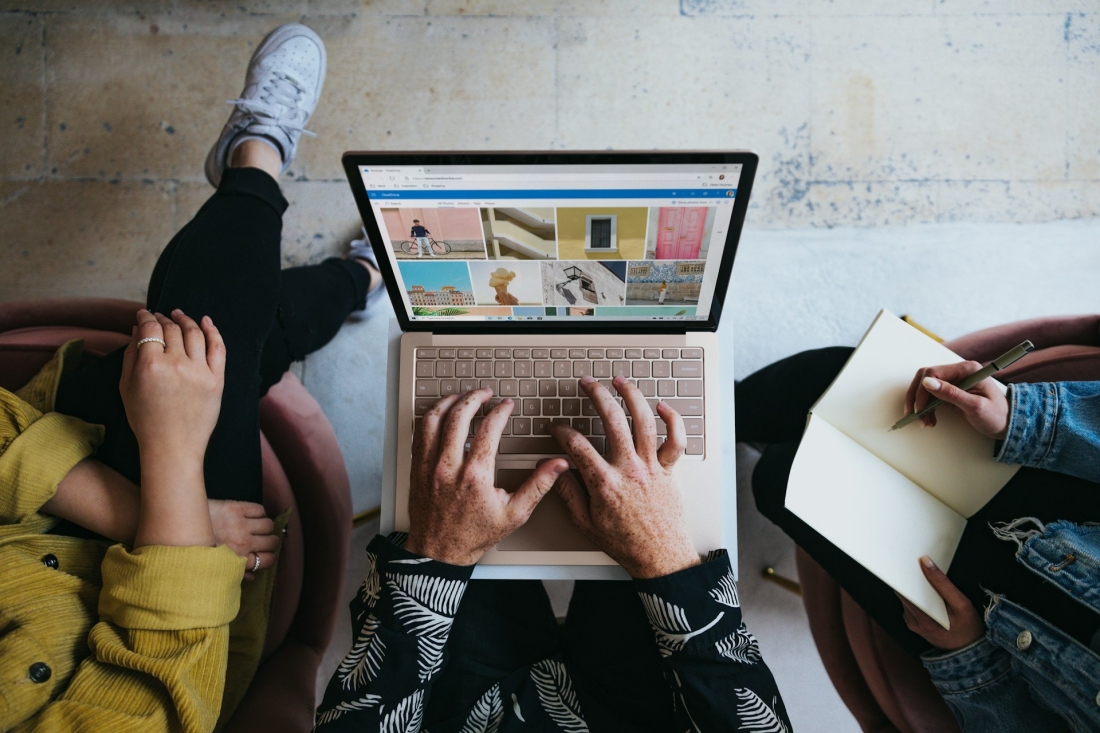 Ansicht von oben: Drei Personen arbeiten gemeinsam. Eine tippt auf einem Laptop mit Bildübersicht, eine macht handschriftliche Notizen.