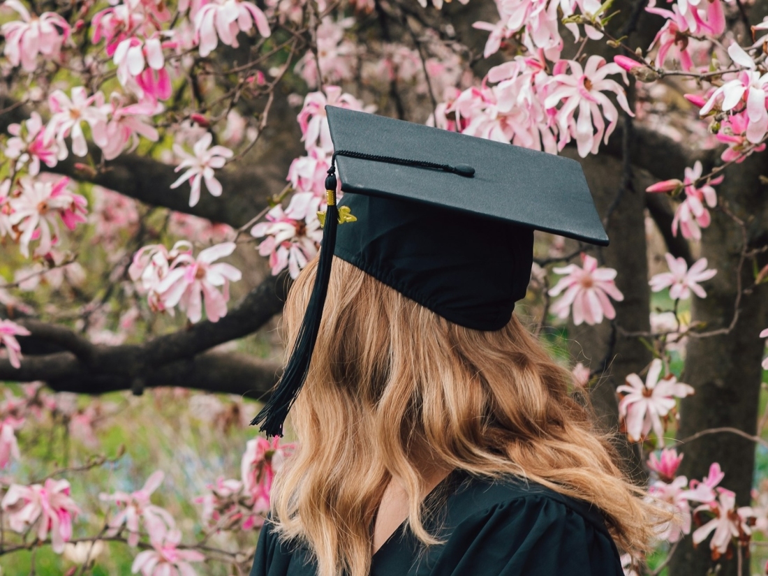 Frau mit langen Haaren im schwarzen Talar vor blühendem Baum trägt Absolventenhut