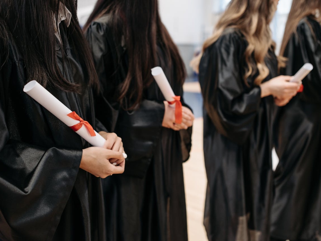 Reihe von Absolventinnen im Talar, jeweils mit gerollter Urkunde mit rotem Band