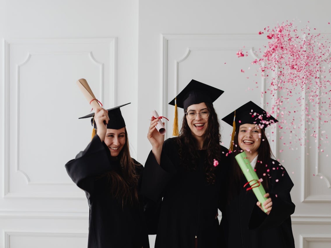 Drei Absolventinnen halten Abschlussurkunden und tragen Absolventenkappen