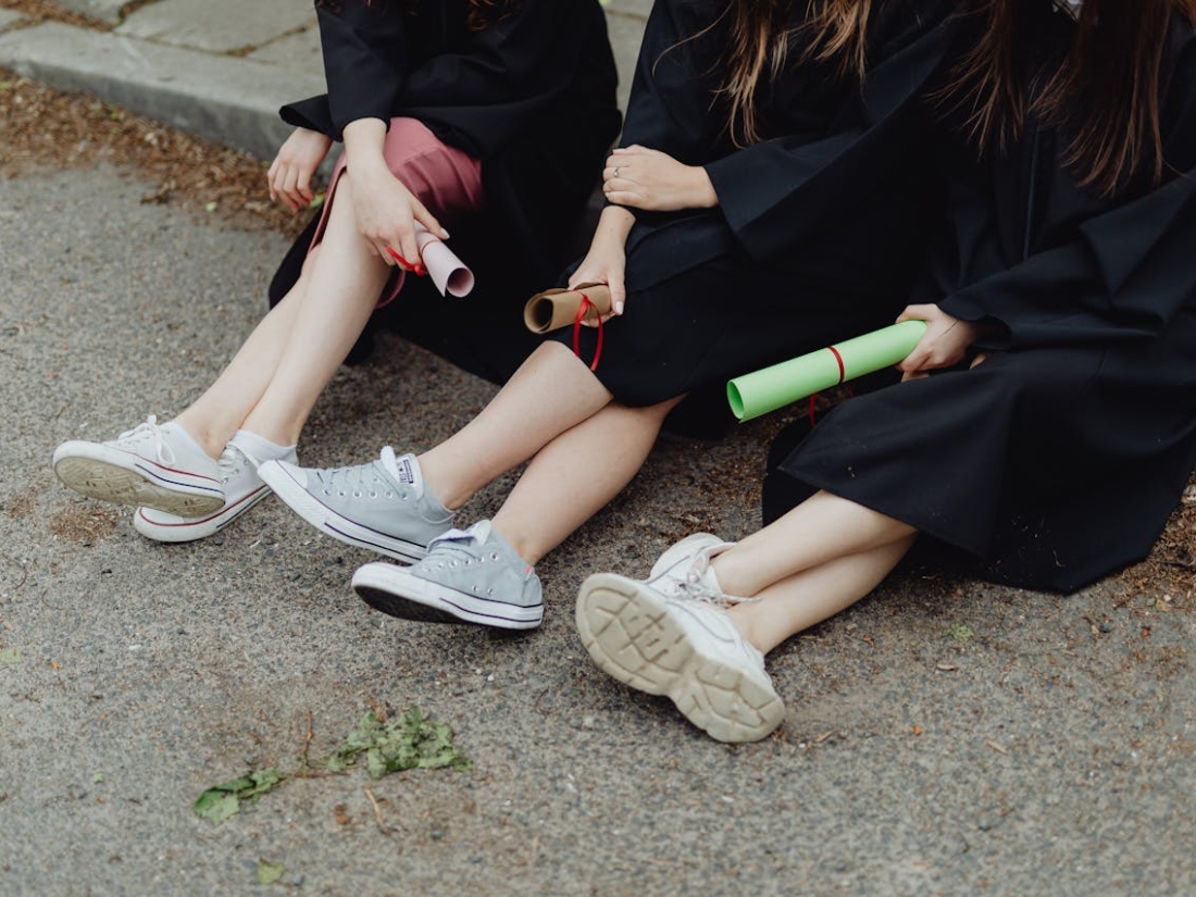 Drei Personen in Abschlusskleidung sitzen am Boden und halten gerollte Urkunden in der Hand.