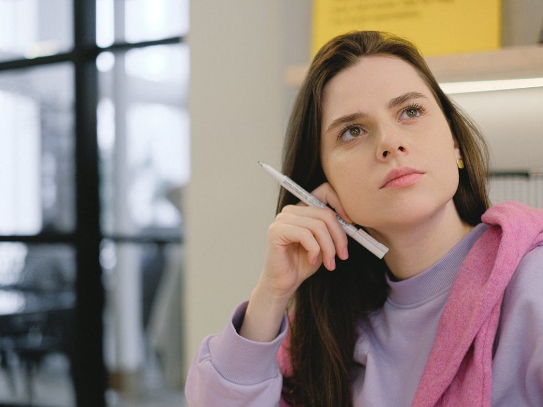 Junge Frau hält einen Stift und blickt nach oben, im Hintergrund ein Büro oder Klassenraum.