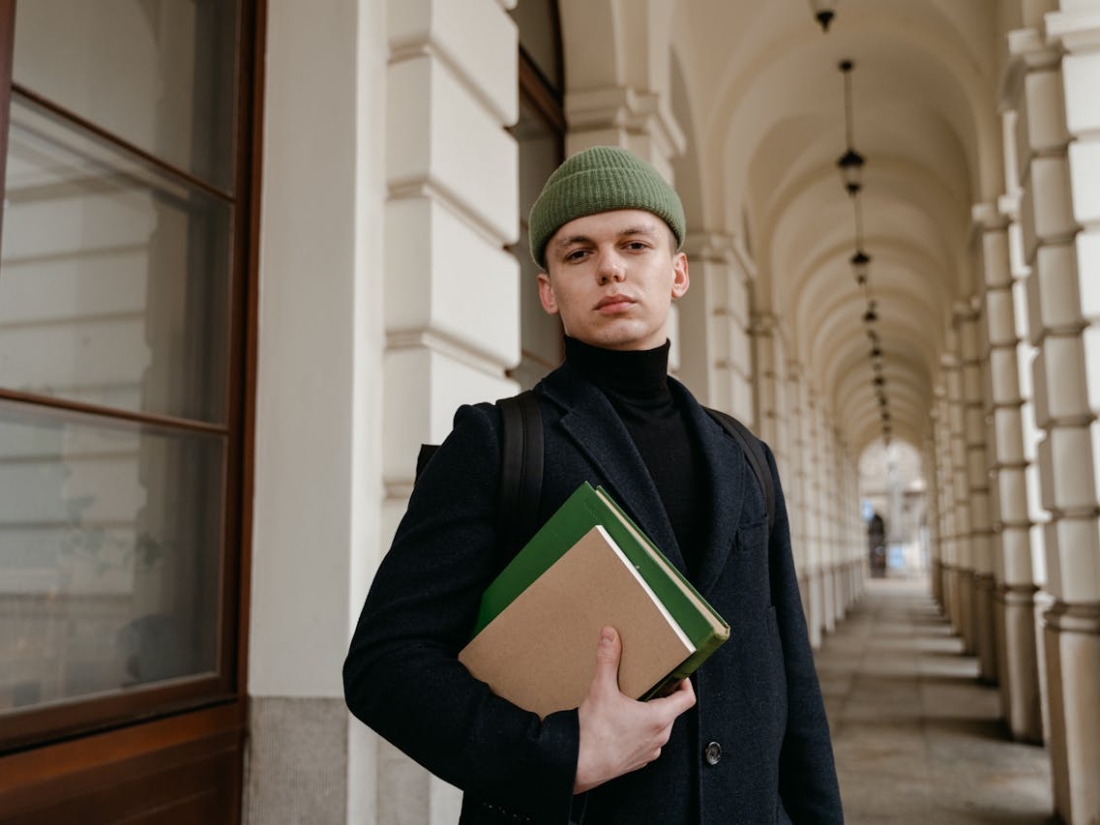 Junger Mann mit Mütze hält Ordner und steht in einem Arkadengang vor einem Gebäude.