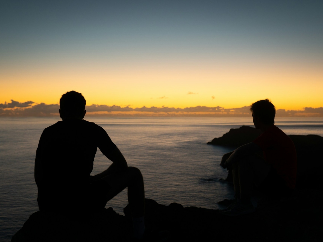 Zwei Personen als Silhouett e sitzen auf Felsen am Meer bei Sonnenuntergang.