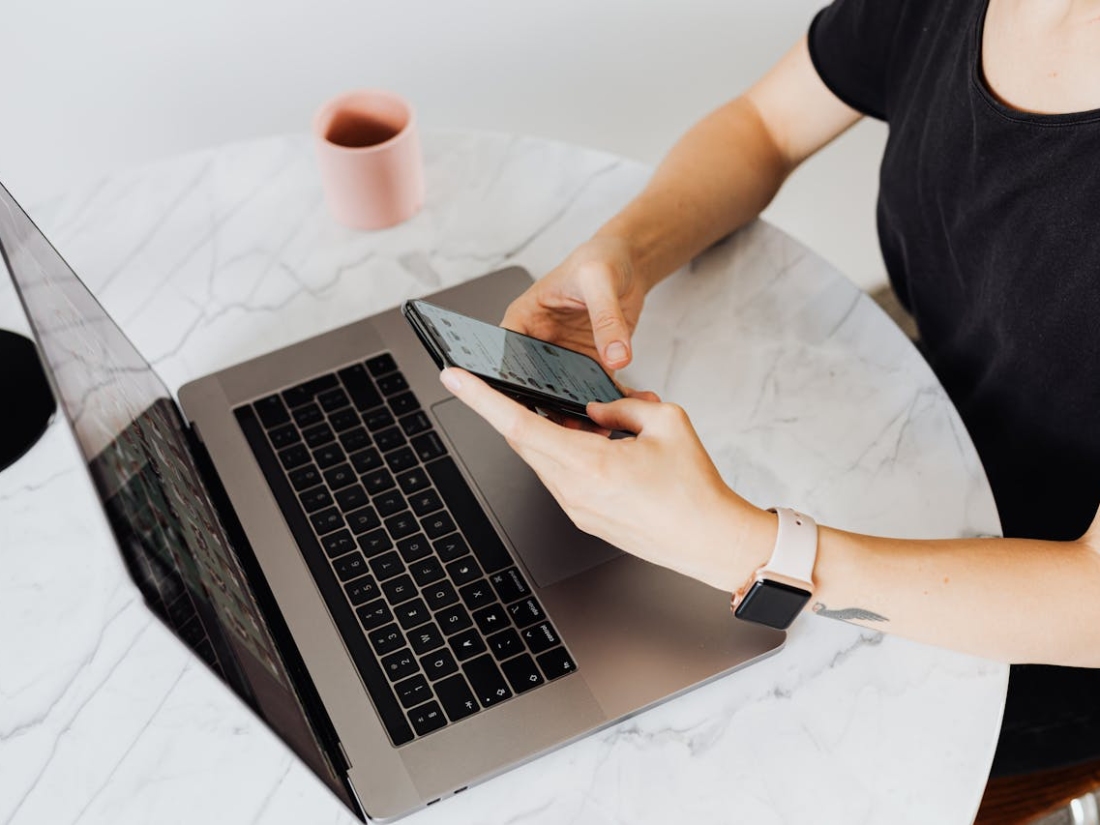 Perspektive auf die Arme einer Person mit Handy in der Hand und Laptop auf dem Tisch.