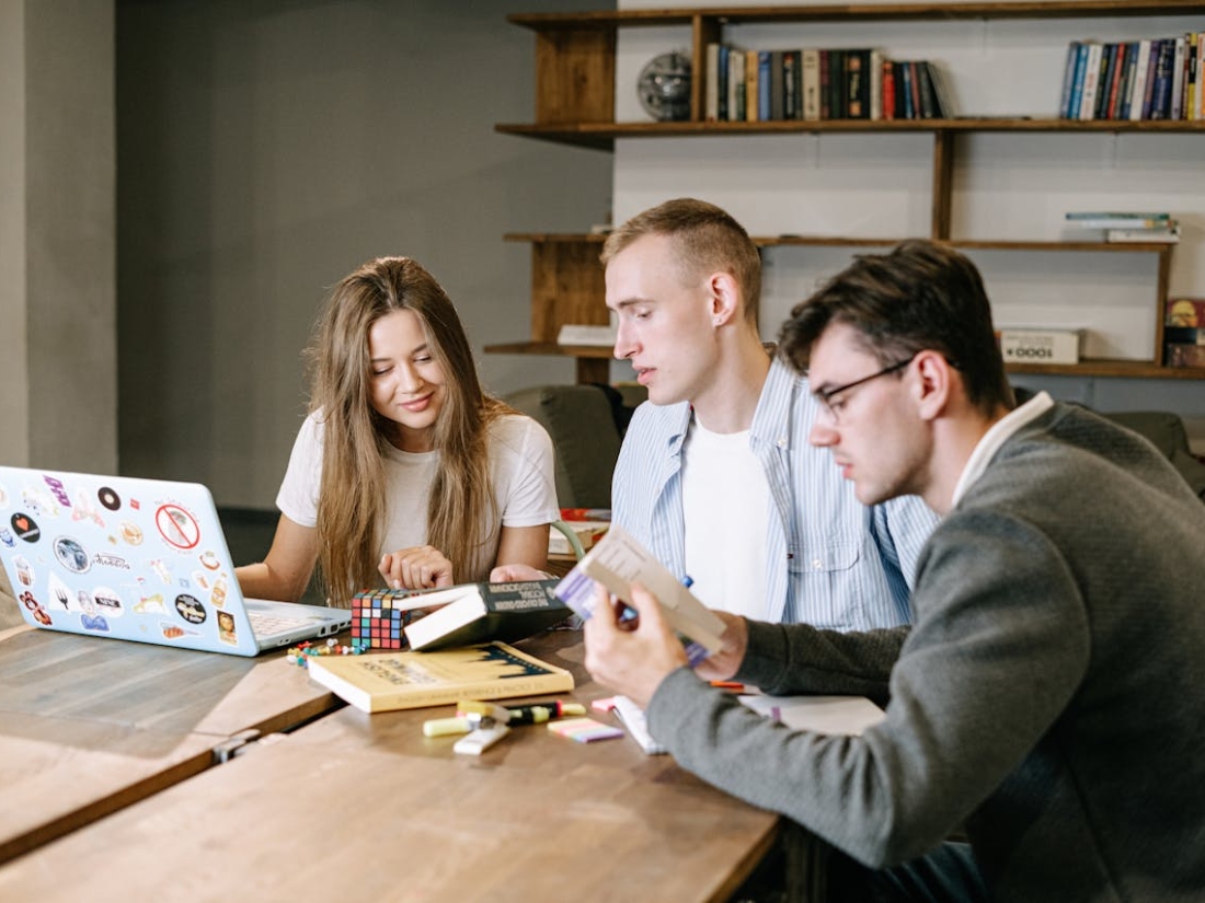 Drei Studierende sitzen an einem Holztisch mit Laptop, Büchern und Notizen und arbeiten gemeinsam an einem Projekt.