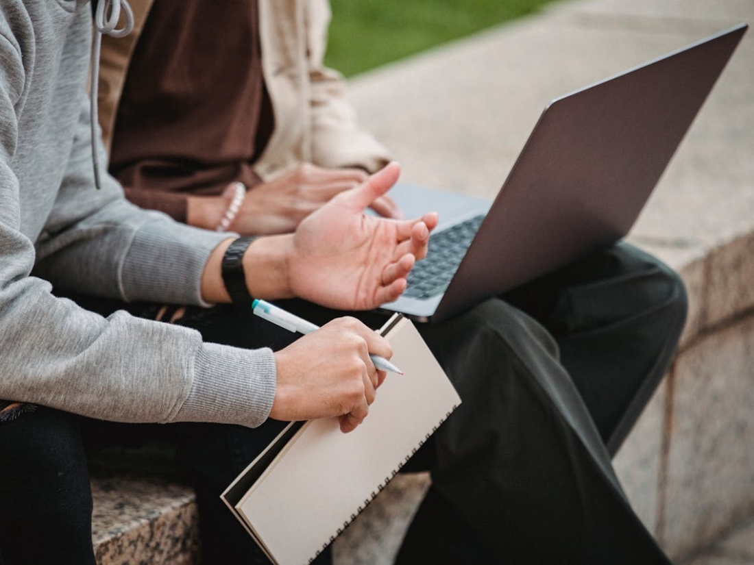 Zwei Personen sitzen auf einer Treppe, eine schreibt in ein Notizbuch, die andere zeigt etwas auf dem Laptop.