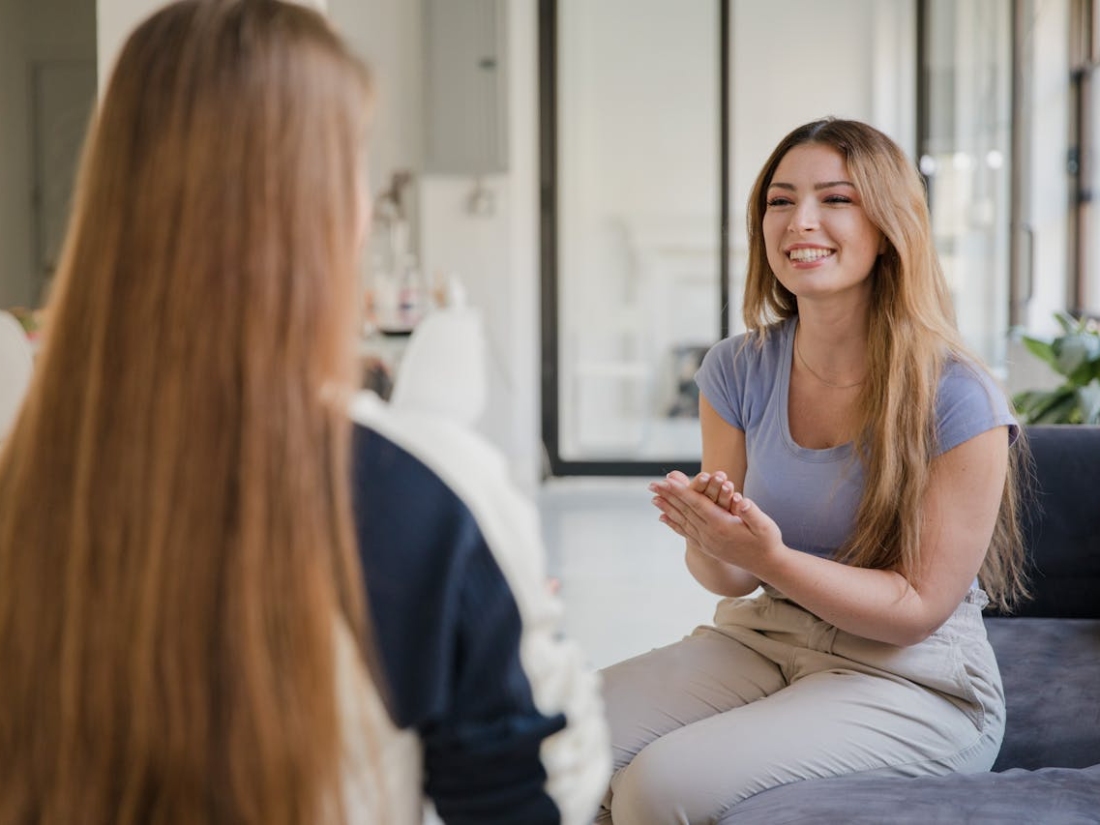 Zwei junge Frauen sitzen sich gegenüber und führen ein entspanntes Gespräch in einem hellen, modernen Raum.