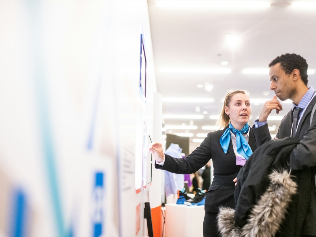 Frau und Mann unterhalten sich an einer Infowand – Master Sportmanagement