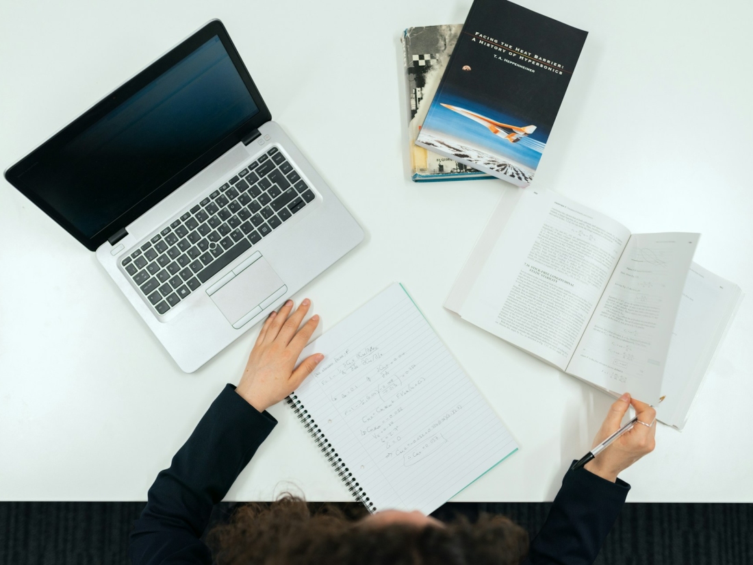 Person sitzt mit Block, Büchern und Stift vor einem Laptop